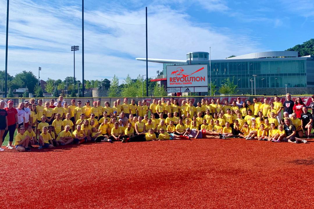 Large group of youth athletes in yellow shirts at a softball camp, posing on a red field with coaches.