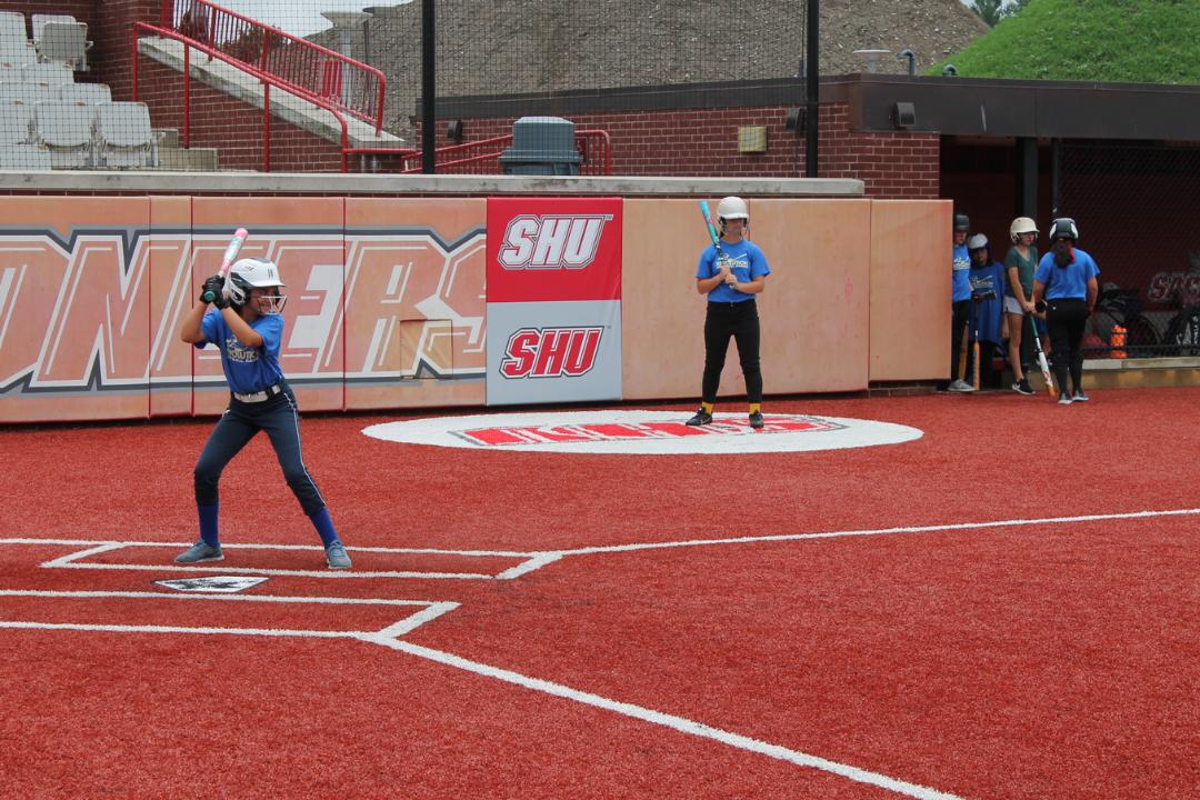 Youth softball players practicing on a red turf field, one batting and others observing.